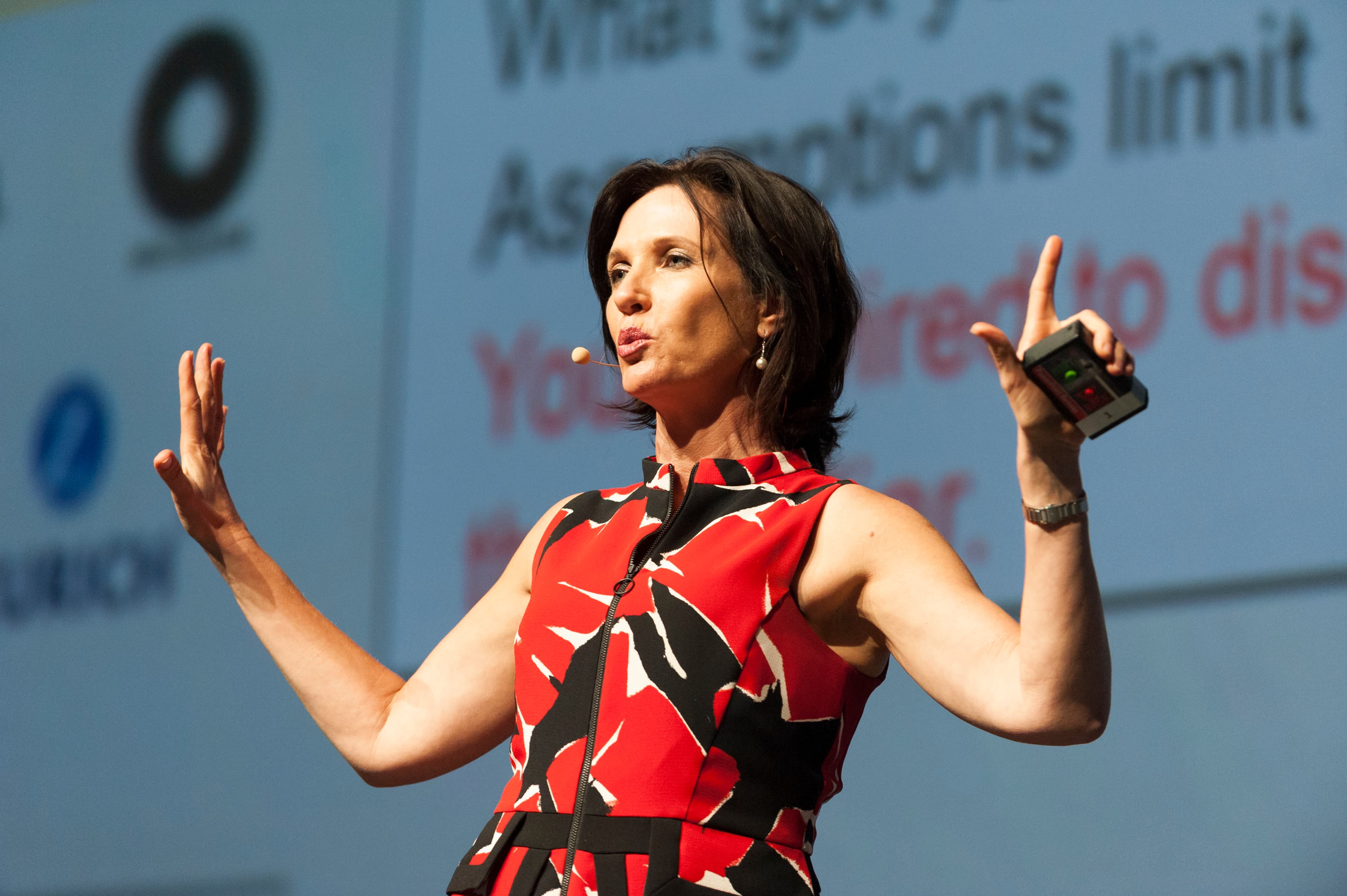 Dr. Margie Warrell mid-keynote in a red and black patterned sleeveless dress, wearing a headset microphone with both hands raised and a presenter clicker in one of them, a large out-of-focus slide behind her